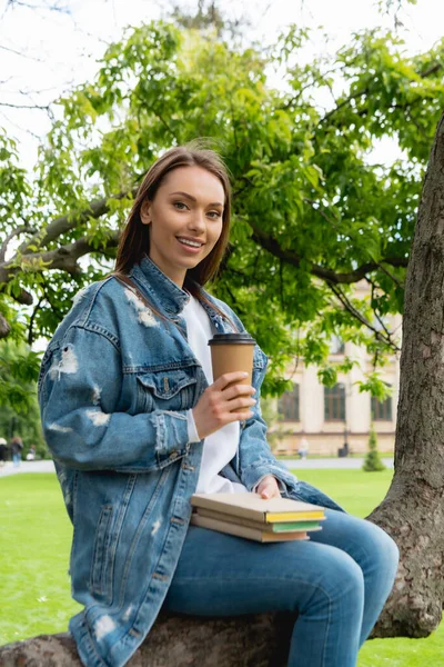 Cheerful and beautiful student holding paper cup and books while sitting on tree — Stock Photo