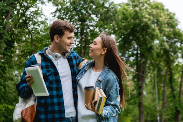 Fröhliche und junge Studentin schaut Mädchen mit Büchern und Pappbecher an — Stockfoto