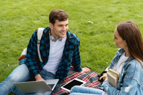Estudiante feliz mirando chica atractiva cerca de gadgets en manta a cuadros — Stock Photo