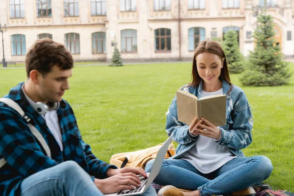 Селективное внимание веселой девушки чтение книги рядом с красивым студентом с помощью ноутбука, концепция онлайн исследования — стоковое фото