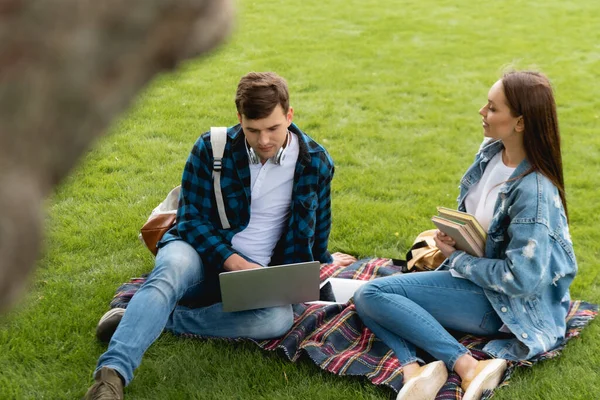 Enfoque selectivo de niña alegre sosteniendo libros y mirando a un estudiante guapo usando el ordenador portátil, concepto de estudio en línea - foto de stock