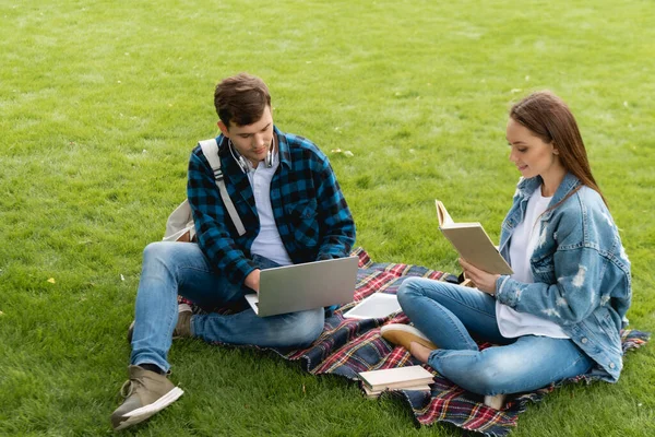 Alegre chica leyendo libro cerca de guapo estudiante usando portátil, concepto de estudio en línea - foto de stock