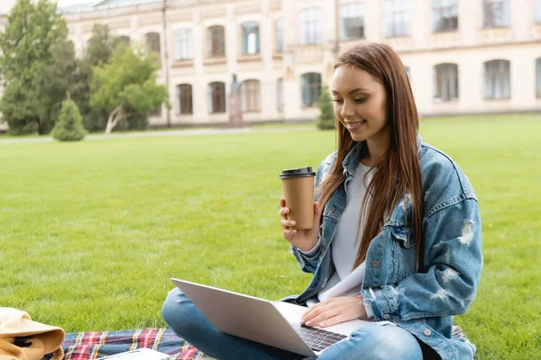 Estudiante feliz y atractivo sosteniendo taza desechable mientras se utiliza el ordenador portátil, concepto de estudio en línea - foto de stock