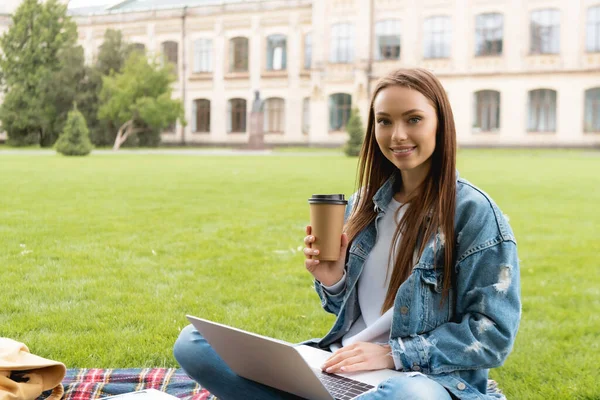 Attrayant étudiant tenant tasse jetable et souriant tout en utilisant un ordinateur portable, concept d'étude en ligne — Photo de stock