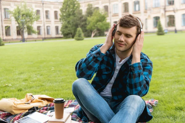 Estudiante feliz con los ojos cerrados tocando auriculares inalámbricos y escuchar música cerca de la computadora portátil y taza de papel en manta a cuadros - foto de stock