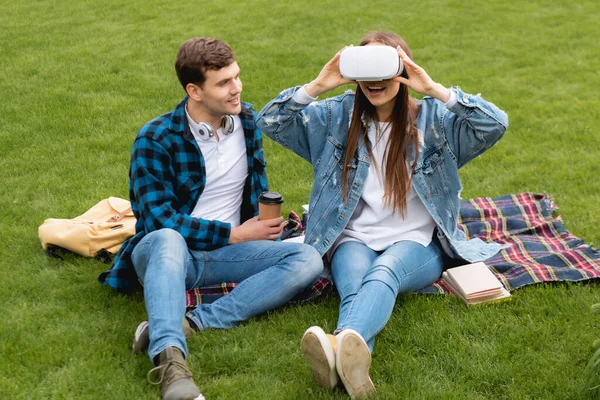 Estudante alegre segurando copo de papel e olhando para menina animado em fone de ouvido realidade virtual — Fotografia de Stock