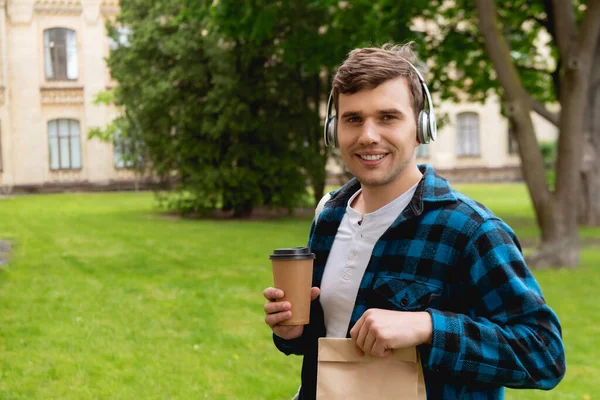 Étudiant joyeux dans les écouteurs sans fil écouter de la musique et tenant sac en papier et café à emporter — Photo de stock