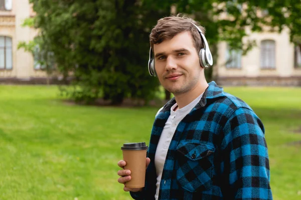 Étudiant joyeux dans les écouteurs sans fil écouter de la musique et tenant tasse en papier — Photo de stock