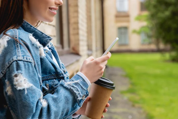 Vista recortada de estudiante alegre usando teléfono inteligente y sosteniendo taza de papel cerca del campus universitario - foto de stock
