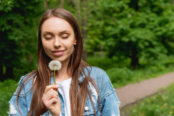 Cheerful young woman with closed eyes holding dandelion in park — Stock Photo