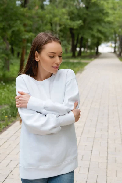 Attraktive junge Frau im Sweatshirt steht mit verschränkten Armen im Park — Stockfoto