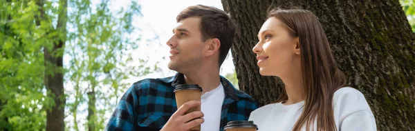 Panoramic orientation of happy students holding paper cups and looking away in green park — Stock Photo