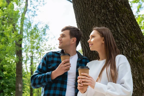 Happy couple holding paper cups and looking away in green park — Stock Photo