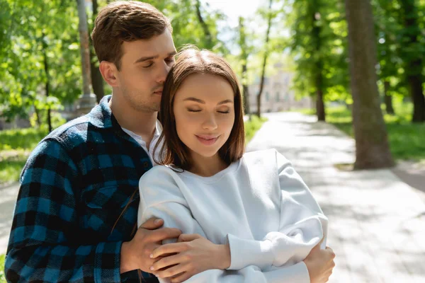 Feliz estudiante abrazando alegre novia con los ojos cerrados en verde parque - foto de stock