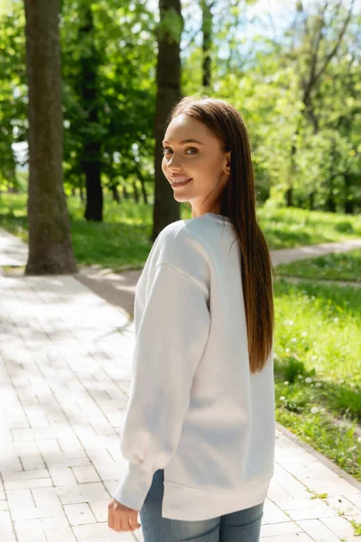 Chica alegre en sudadera blanca de pie en el parque y mirando a la cámara - foto de stock
