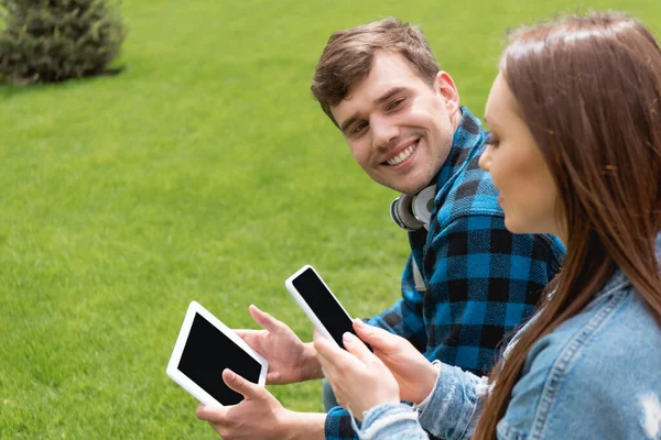 Вибірковий фокус веселого студента, який дивиться на дівчину зі смартфоном, тримаючи цифровий планшет з порожнім екраном — стокове фото