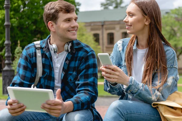 Estudiante feliz y atractiva chica sosteniendo gadgets y mirándose entre sí - foto de stock