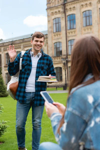 Enfoque selectivo de estudiante guapo sonriendo mientras sostiene libros y agitando la mano cerca de la chica con el teléfono inteligente - foto de stock