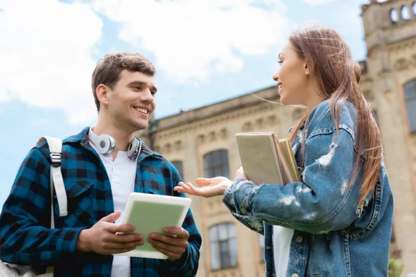 Low angle view of cheerful girl gesturing while holding books and talking with happy student — Stock Photo