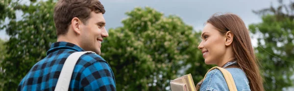 Panoramic crop of happy and young woman holding books and looking at handsome student — Stock Photo