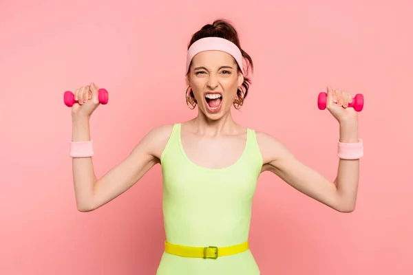 Emotionale Sportlerin blickt in die Kamera und trainiert mit Hanteln auf rosa — Stockfoto