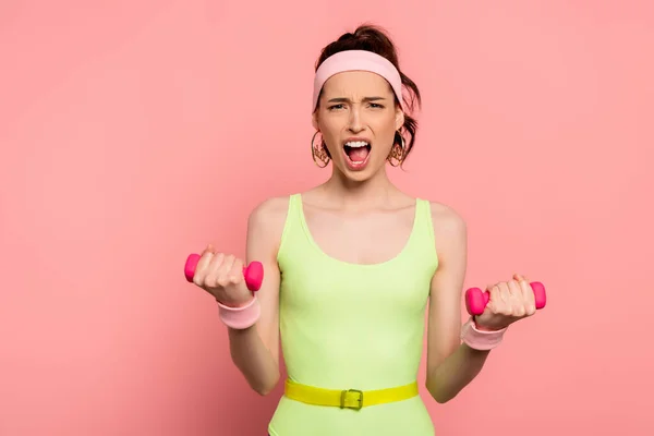 Chica emocional mirando a la cámara y haciendo ejercicio con mancuernas en rosa - foto de stock