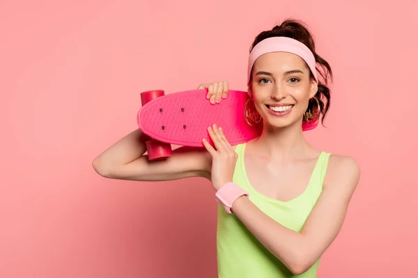 Cheerful girl with headband holding penny board on pink — Stock Photo
