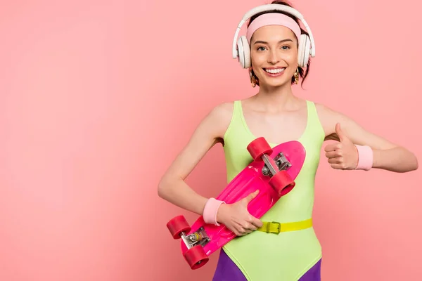 Happy girl in bodysuit and headphones showing thumb up while holding penny board on pink — Stock Photo