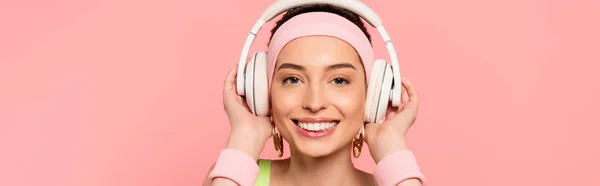 Imagen horizontal de chica alegre tocando auriculares mientras escucha música aislada en rosa - foto de stock