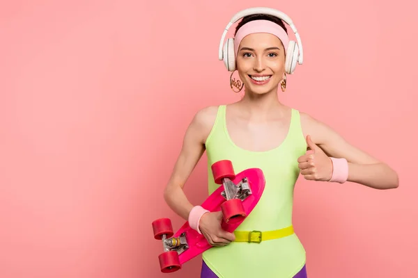 Cheerful girl in bodysuit and headphones showing thumb up while holding penny board on pink — Stock Photo