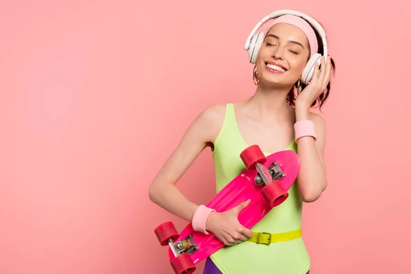 Cheerful girl in bodysuit touching headphones while holding penny board isolated on pink — Stock Photo
