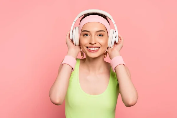 Весела дівчина торкається навушників під час прослуховування музики ізольовано на рожевому — стокове фото