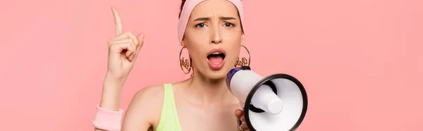 Horizontal concept of emotional young woman pointing with finger and holding loudspeaker isolated on pink — Stock Photo