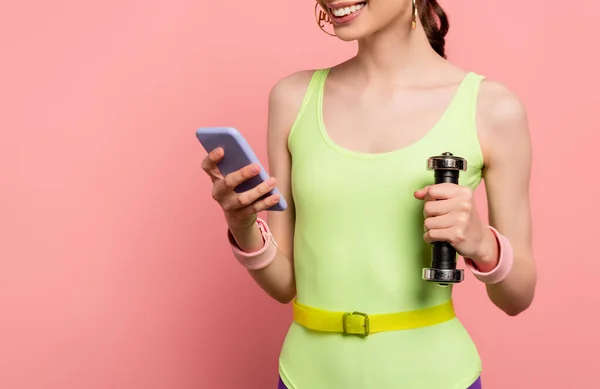 Обрезанный вид веселой спортсменки держа гантель и используя смартфон на розовый — стоковое фото