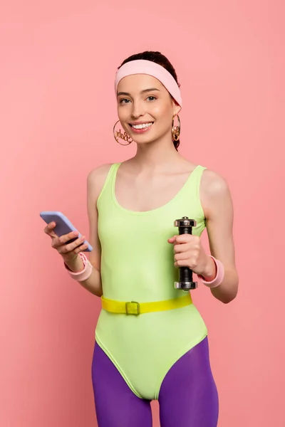 Cheerful sportswoman holding dumbbell and using smartphone isolated on pink — Stock Photo