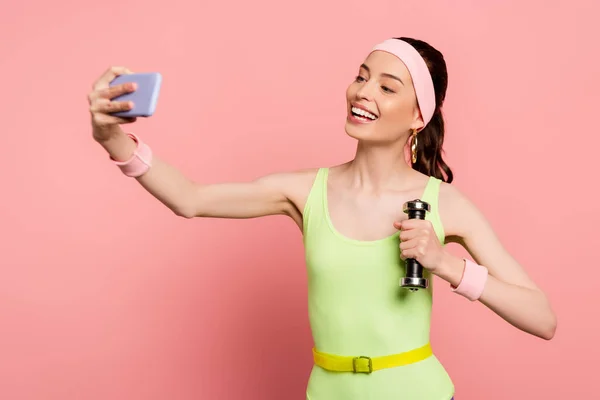 Cheerful sportswoman holding dumbbell and taking selfie on pink — Stock Photo