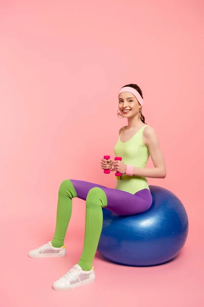 Sportive heureuse assise sur une balle de fitness et faisant de l'exercice avec des haltères sur rose — Photo de stock