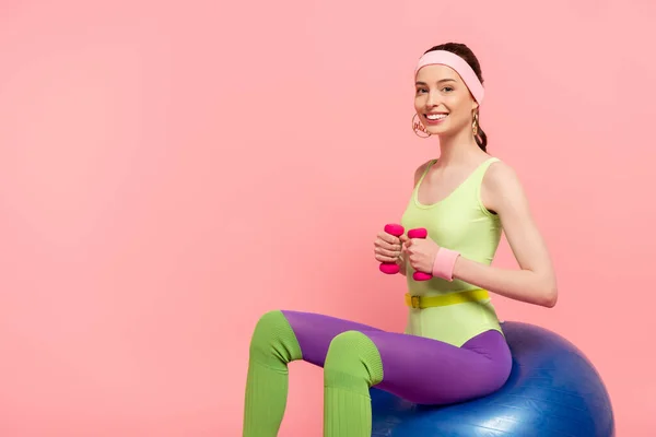 Mujer feliz sentada en la pelota de fitness y haciendo ejercicio con pesas aisladas en rosa — Stock Photo