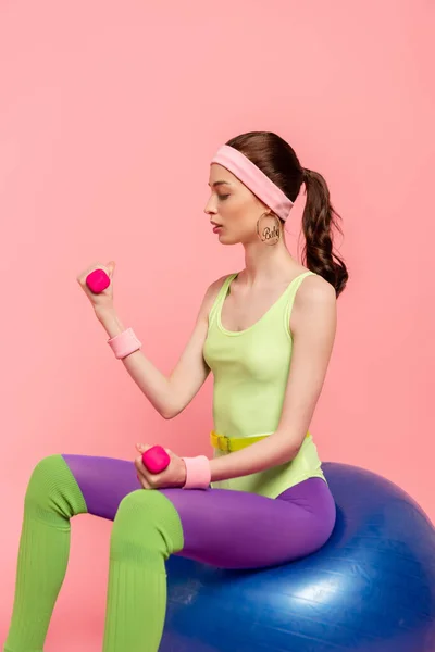 Jovem desportista sentado na bola fitness e exercitando-se com halteres isolados em rosa — Fotografia de Stock