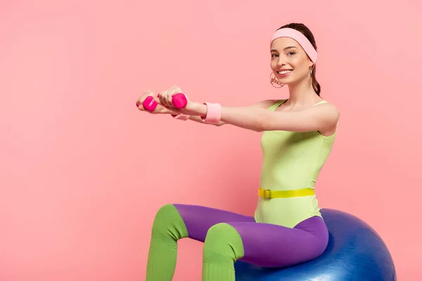 Deportista sonriente con las manos extendidas haciendo ejercicio con pesas y sentado en la pelota de fitness en rosa - foto de stock