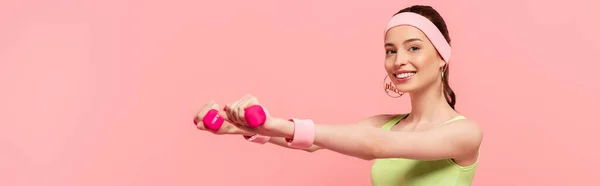Panoramaaufnahme einer lächelnden Sportlerin mit ausgestreckten Händen, die mit auf rosa isolierten Hanteln trainiert — Stockfoto