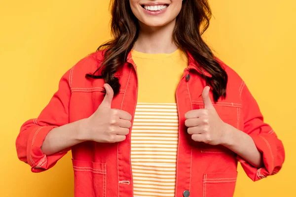 Обрезанный вид веселой молодой женщины, показывающей большие пальцы вверх и смотрящей в камеру на желтом — стоковое фото