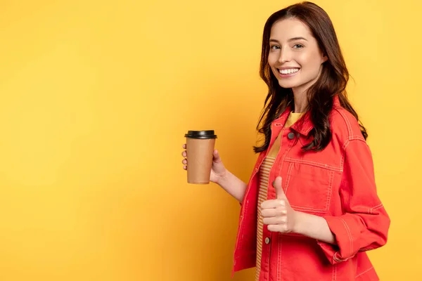Весела молода жінка показує великий палець і тримає паперову чашку на жовтому — стокове фото