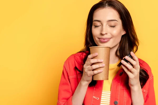 Fröhliche junge Frau hält Pappbecher in der Hand und riecht Kaffee to go auf gelb — Stockfoto