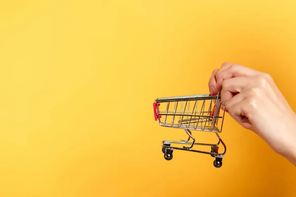 Vista cortada de mulher segurando carrinho de compras pequeno no amarelo — Fotografia de Stock