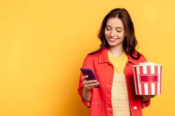 Happy woman holding popcorn bucket and using smartphone on yellow — Stock Photo