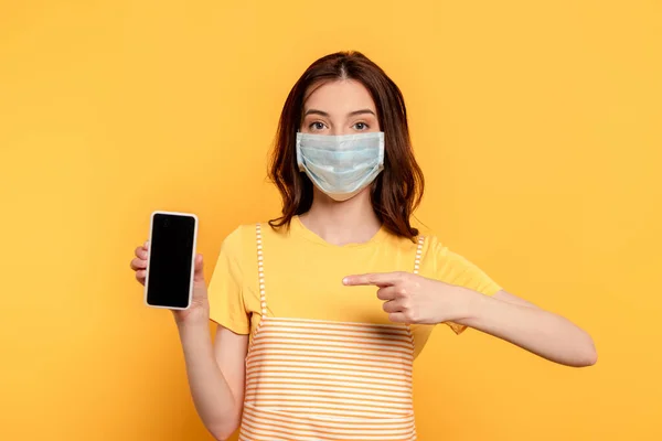 Junge Frau in medizinischer Maske zeigt mit dem Finger auf Smartphone mit leerem Bildschirm auf gelb — Stockfoto