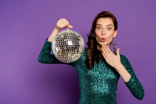 Surprised young woman in dress holding disco ball on purple — Stock Photo
