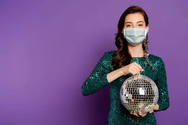 Jovem mulher em vestido e máscara médica segurando bola de discoteca em roxo — Fotografia de Stock