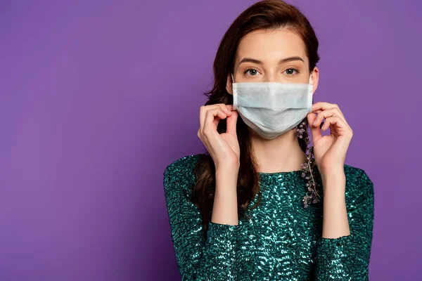 Mujer joven en vestido tocando máscara médica y mirando a la cámara en púrpura - foto de stock